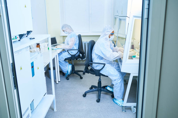 Vista lateral completa de dos científicos que usan trajes de protección y máscaras que trabajan en mesas opuestas en el laboratorio de investigación
 - Foto, Imagen
