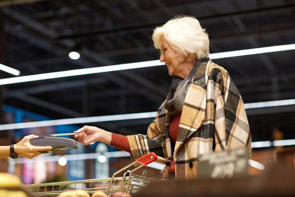 Side view portrait of modern senior woman paying with NFC payment via smartphone  and smiling happily while buying groceries in supermarket - Photo, Image
