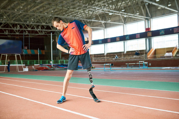 Full length portrait of young handicapped sportsman with artificial foot warming up before running practice in modern gym stretching to side - Photo, Image