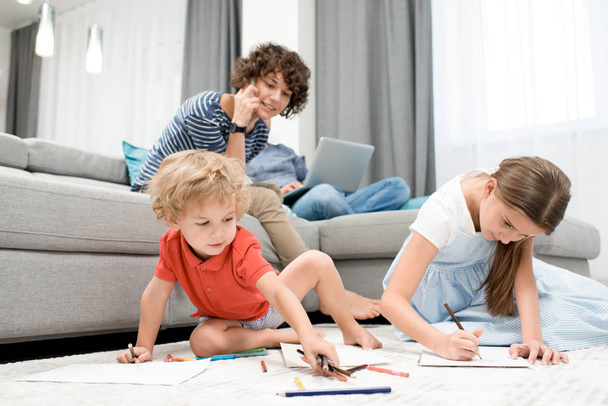 Portret dwojga dzieci, rysunek zdjęcia, siedząc na dywanie w salonie z Mama, obserwując je z sofą - Zdjęcie, obraz