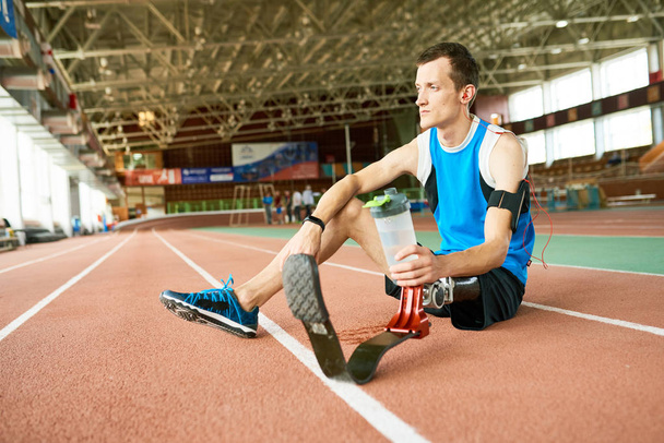Retrato de atleta amputado pensivo sentado na pista de corrida fazendo uma pausa da prática para relaxar e beber água
 - Foto, Imagem