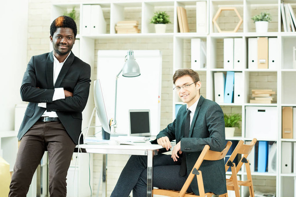 Retrato de grupo de jóvenes gerentes financieros guapos que usan trajes mirando a la cámara con sonrisas anchas mientras tienen reunión de trabajo en una oficina abierta
 - Foto, imagen