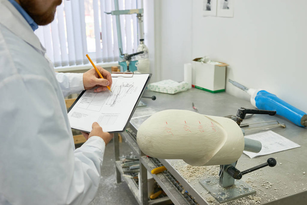 Retrato del joven técnico de prótesis escribiendo notas en el portapapeles mientras comprueba el molde de prótesis en el laboratorio de diseño
 - Foto, imagen