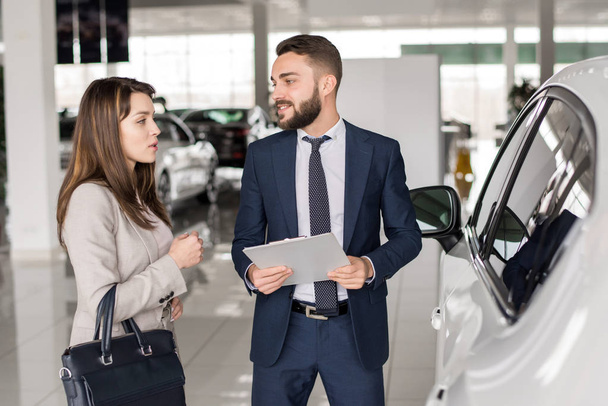 Retrato de vendedor de carro bonito falando com a jovem mulher ajudando-a a escolher o carro de luxo no showroom
 - Foto, Imagem