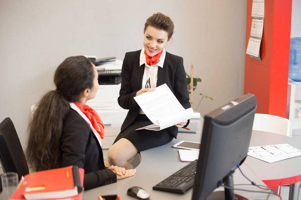 Ritratto di giovane consulente aziendale in uniforme nera e rossa seduto sulla scrivania e sorridente mentre mostra i documenti al collega nell'ufficio moderno
 - Foto, immagini