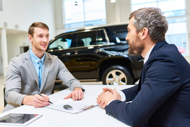 Портрет успешного молодого человека, покупающего новый роскошный автомобиль в выставочном зале, подписывающего договор купли-продажи, сидя напротив зрелого продавца
 - Фото, изображение