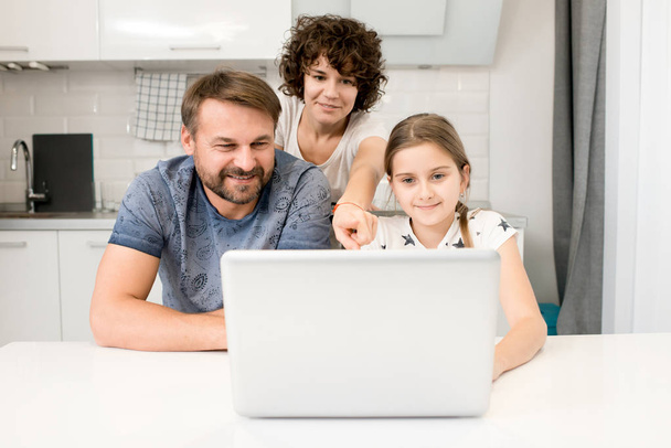 Portrait de jeune famille heureuse regardant des vidéos via un ordinateur portable assis à la table de cuisine dans un appartement moderne
 - Photo, image