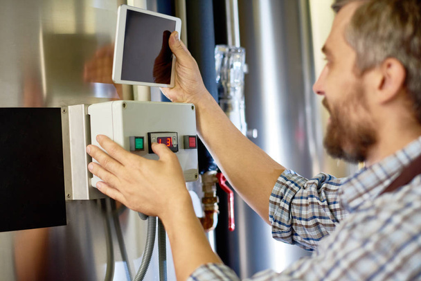 Close-up of concentrated brewing worker viewing maintenance instruction on digital tablet while adjusting brewing equipment at modern beer factory - Foto, immagini