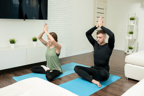 Retrato de pareja joven moderna haciendo ejercicios de yoga en la sala de estar en casa, meditando en posición de loto con los ojos cerrados
 - Foto, Imagen