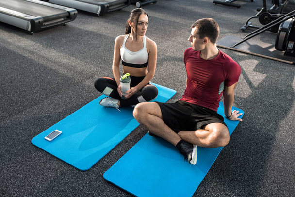 Retrato de alto ángulo de una joven pareja deportiva tomando un descanso mientras hace yoga sobre alfombras en el gimnasio moderno
 - Foto, Imagen