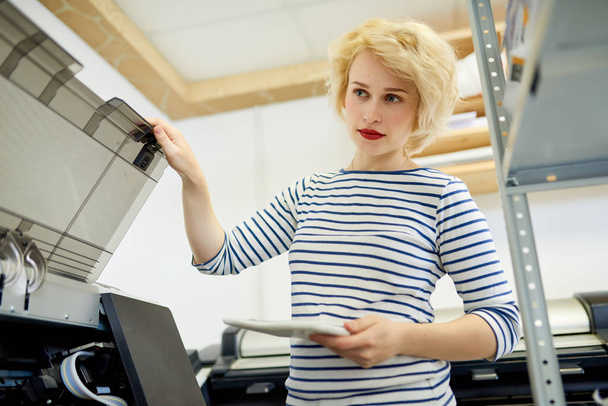 Low angle portrait of blonde young woman closing plotter or copy machine while working in printing shop - Fotoğraf, Görsel