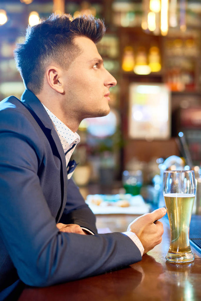 Zobacz Profil przystojny młody człowiek sobie stylowy garnitur siedzi licznik bar po ciężkim dniu pracy, picia piwa i oglądając mecz rugby - Zdjęcie, obraz