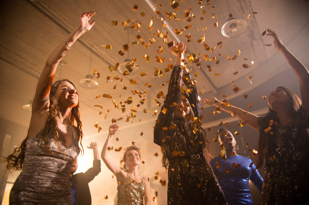Grupo de belas mulheres jovens dançando sob chuveiro de confete dourado desfrutando de festa delirante em boate, baixo ângulo
 - Foto, Imagem