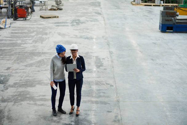 Full length portrait of two modern female engineers wearing hardhats planning work writing on clipboard in workshop of modern factory - Фото, изображение