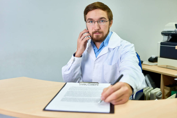 Portrét přátelské vousatý doktor předání pen a kontrakt na pacienta umístění na stole v kanceláři a používat smartphone  - Fotografie, Obrázek