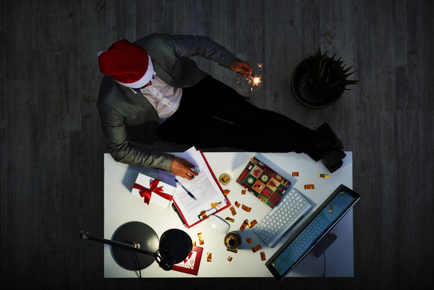 Celebración de la Nochebuena en el trabajo: trabajador irreconocible de cuello blanco con sombrero de Santa sentado con piernas en el escritorio y con chispa de fuegos artificiales en la mano, interior de la oficina oscura moderna
 - Foto, imagen
