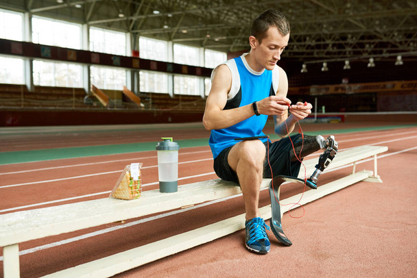 Retrato de comprimento total do jovem atleta amputado fazendo uma pausa da prática na pista de corrida e relaxando no banco
 - Foto, Imagem