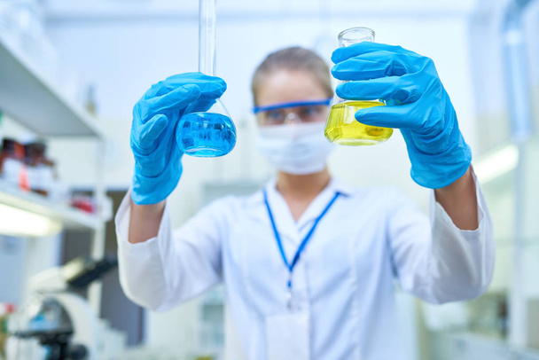 Portret van vrouwelijke wetenschapper blauwe beschermende handschoenen, reageerbuisjes met gekleurde vloeistoffen te houden tijdens de studie van chemische stoffen in medisch laboratorium, focus op handen dragen - Foto, afbeelding