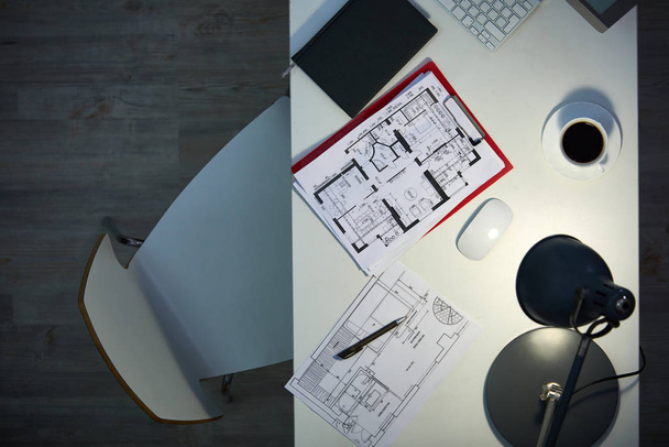 Working late in modern design studio: directly above view of wooden desk with floor plans and cup of coffee illuminated with task lamp, no people - Photo, Image