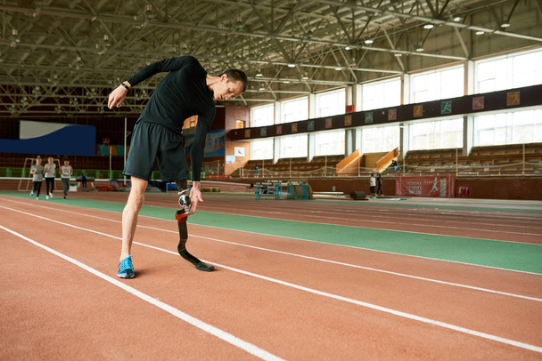 Ganzkörperporträt eines jungen behinderten Athleten mit künstlichem Fuß beim Aufwärmen vor dem Lauftraining in einer modernen Turnhalle, die sich seitlich streckt - Foto, Bild