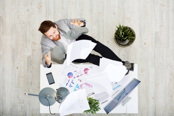 Cheerful bearded entrepreneur sitting at office desk and throwing documents in air after successful completion of promising project, directly above view - Photo, Image