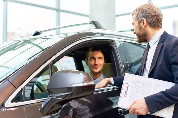 Portrait of handsome young man taking luxury car for test drive, sitting inside and smiling at sales manager in showroom - Photo, Image
