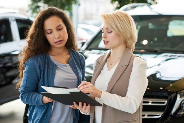 Attractive young consultant discussing contract details with mixed race customer before signing it after successful deal in car showroom - Foto, immagini