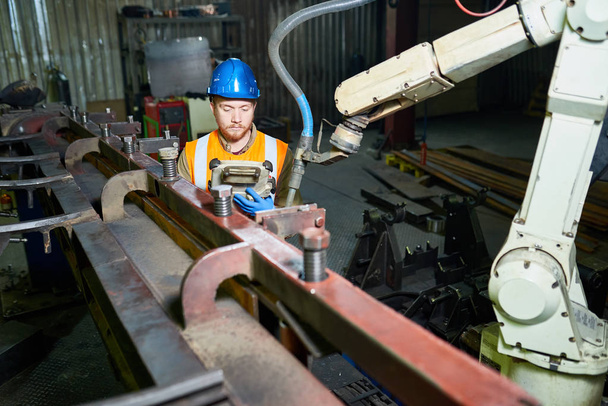 Kopf und Schultern Porträt eines bärtigen rothaarigen Technikers mit reflektierender Weste und harter Arbeitsmaschine, im Hintergrund das Innere der Produktionsabteilung - Foto, Bild