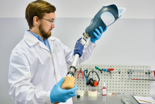 Портрет молодого техника протеза, держащего протез ноги, проверяющего его на качество и выполняющего регулировку во время работы в современной лаборатории
 - Фото, изображение