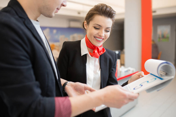 Retrato de bela jovem mulher vestindo uniforme preto e vermelho conversando com o cliente em banco segurando prancheta
 - Foto, Imagem