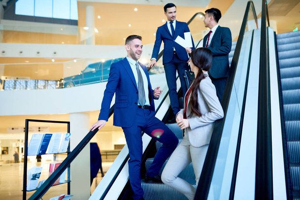 Group of modern young business people riding escalator in office building and chatting cheerfully on the way, copy space - Photo, image