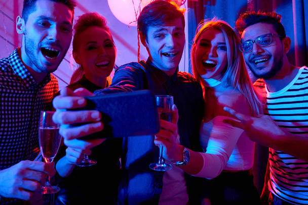 Skupina s úsměvem mladých lidí, kteří selfie vychutnávat sváteční výzdobou v soukromém domě party při pohledu na fotoaparát smartphonu - Fotografie, Obrázek