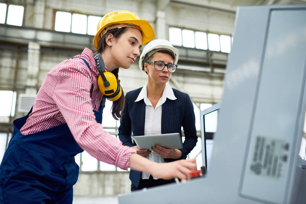 Retrato de ángulo bajo de dos mujeres jóvenes que usan cascos duros que trabajan en una planta moderna junto al panel de control de la unidad de la máquina
 - Foto, Imagen
