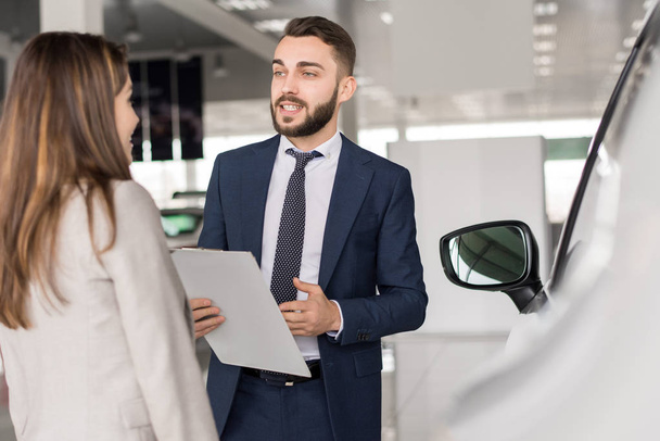 Portrait of handsome car salesman talking to young woman helping her choose in car showroom - Photo, Image