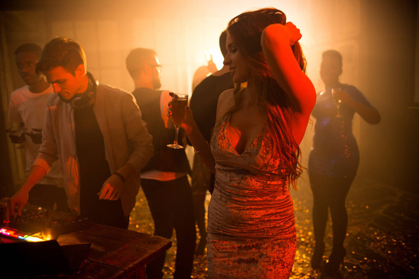 Porträt eines schönen jungen Mädchens, das elegant inmitten einer schummrigen Tanzfläche tanzt, Champagnerglas in der Hand hält und ein modisches Samtkleid trägt - Foto, Bild