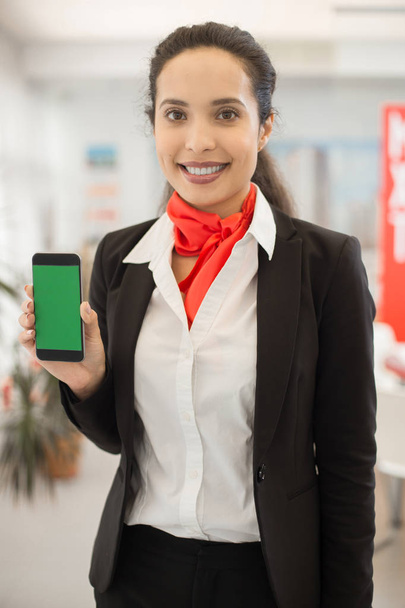 Taille portret van gemengd ras vrouw die lacht op camera houden smartphone met lege groen scherm presenteren mobiele app in kantoor - Foto, afbeelding