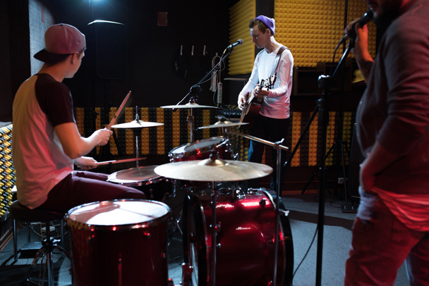 Band of young musicians performing in dim recording studio making new album, drum set in foreground - Photo, Image