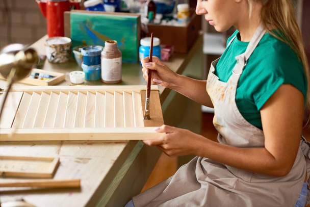 Vista lateral de la joven rubia disfrutando del trabajo en el estudio de arte pintando persianas con pintura de bronce, haciendo decoraciones de interiores de bricolaje
 - Foto, imagen