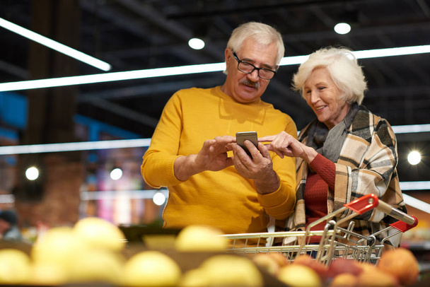 Üst düzey çift bakkal alışveriş okumak için smartphone kullanarak süpermarkette alışveriş portresi listelemek ve meyve ve sebze Isle, kopya alanı tarafından duran fiyatlarını hesapla - Fotoğraf, Görsel