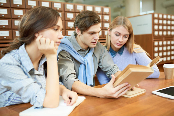 Two pretty students and their handsome groupmate gathered together in spacious library and studying hard, catalogue cabinets on background - Photo, Image