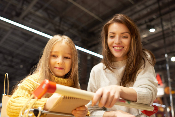 Портрет веселої молодої жінки продовольчі покупки з дочкою в супермаркеті читаючи список покупок і спираючись на візок
 - Фото, зображення