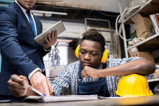 Jistý africký Američan soustruh operátor nosit chrániče sluchu sedí u pracovního stolu a diskutovat výsledky práce s jeho nadřízený, vnitřní katedry produkce na pozadí - Fotografie, Obrázek