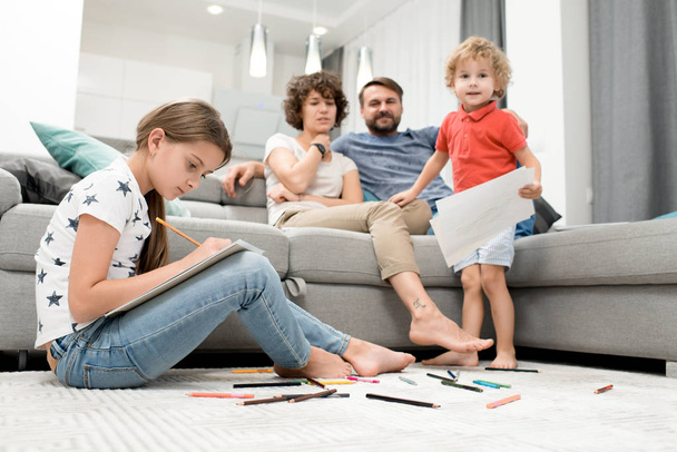 Lovely moments of parenthood: curly middle-aged woman and her bearded husband sitting on cozy sofa and keeping eye on their kids while they drawing colorful pictures with pencils - Fotoğraf, Görsel