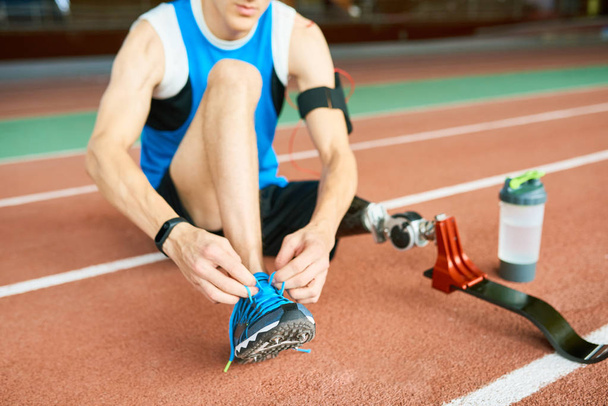 Atleta amputado irreconocible atando cordones de zapato sentado en pista de atletismo en el moderno estadio interior
 - Foto, imagen