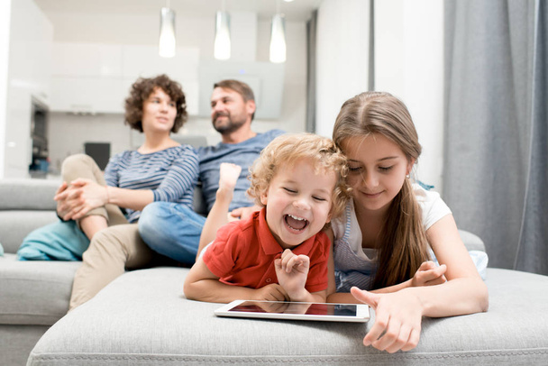 Portrait de frère et sœur utilisant une tablette numérique et riant tout en étant allongé sur le canapé dans le salon avec les parents regardant la télévision en arrière-plan, espace de copie
 - Photo, image