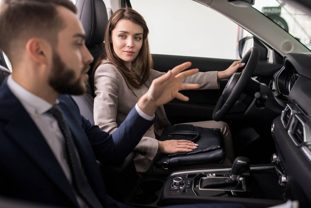 Retrato de bela jovem sentada ao volante do novo carro de luxo e conversando com o vendedor ao escolher o novo transporte
 - Foto, Imagem