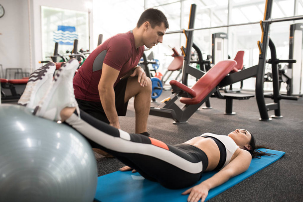 Retrato de cuerpo entero del entrenador de fitness muscular ayudando a la joven a hacer ejercicios en la estera en el gimnasio moderno
 - Foto, imagen