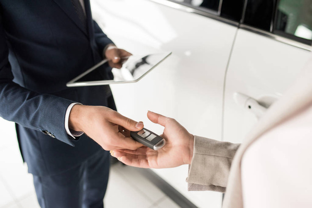Μεγάλη γωνία εσωτερικη του πωλητή αυτοκινήτων δίνοντας κλειδιά υπολογιστή-πελάτη που στέκεται δίπλα σε λευκό πολυτελές αυτοκίνητο - Φωτογραφία, εικόνα