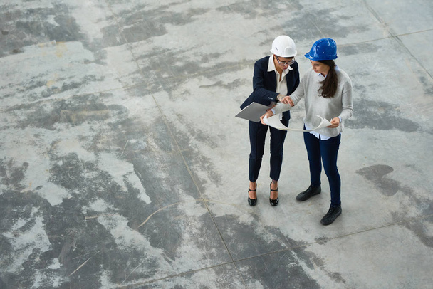 Blick aus der Vogelperspektive auf zwei Ingenieurinnen, die Harthüte tragen und Arbeitspläne in der Werkstatt einer modernen Fabrik untersuchen - Foto, Bild