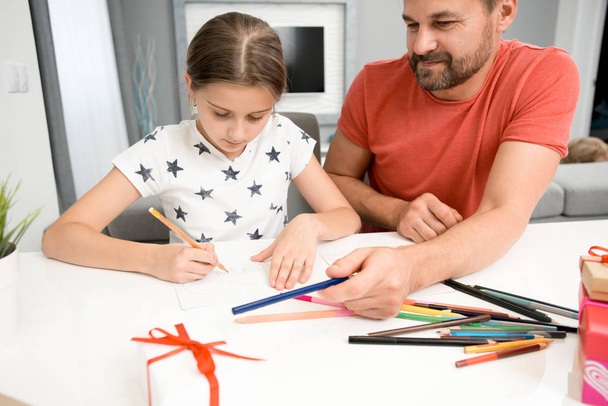 Portrait of bearded father helping little girl drawing handmade card for mothers day at home in modern living room - Photo, Image